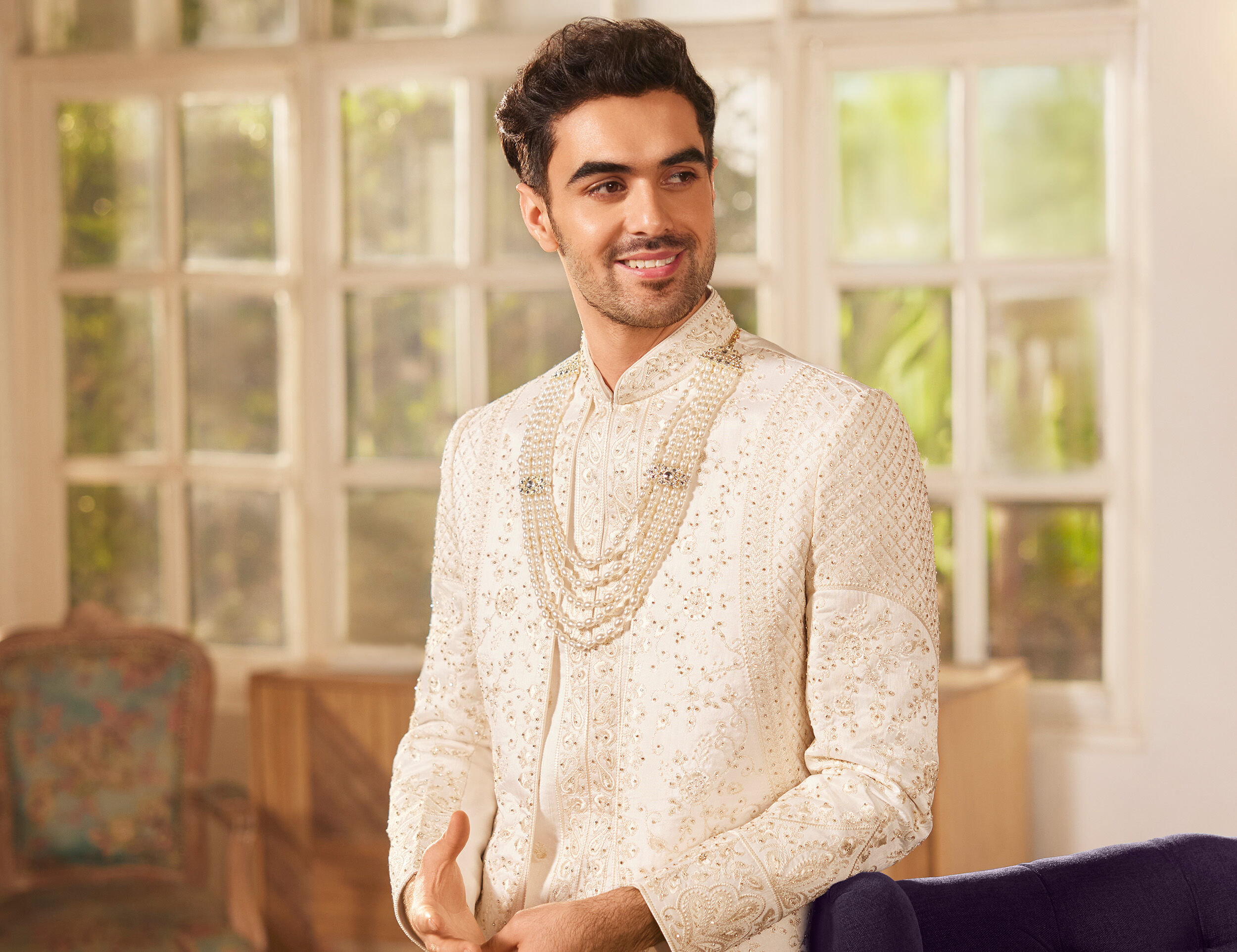 Man in an ivory embroidered sherwani with layered pearl necklace, smiling in a bright, elegant room with large windows 